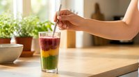 Woman's hand stirring layered drink with straw in bright kitchen, featuring red, yellow and green liquid layers, potted plants and wooden surface for beverage, summer refreshment and healthy lifestyle content.