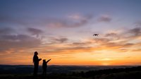 Woman and child silhouettes watching drone fly at sunset over landscape. Aerial technology, family outdoor recreation, and evening sky perfect for travel and leisure content.