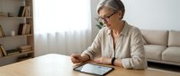 Senior woman with glasses using digital tablet at home sitting at wooden table in modern living room for online communication and technology.