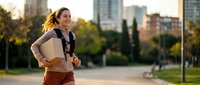 Young professional businesswoman running with laptop bag in urban park during golden hour sunset with city skyline background for active lifestyle concept.