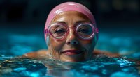 Senior woman in pink swimming cap and goggles smiling while swimming in blue pool water, close-up portrait showing active lifestyle and vitality in older age.