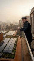 Mature man in dark coat and flat cap standing on urban rooftop with solar panels, overlooking foggy cityscape. Renewable energy and sustainable living concept for commercial use.