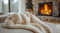 Cozy cream knitted blanket draped on sofa with warm fireplace glowing in background, creating comfortable home atmosphere for relaxation and winter comfort.