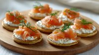 Mini blini pancakes topped with cream cheese, smoked salmon and fresh dill garnish arranged on wooden serving board, elegant appetizer for party catering.