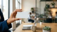 Professional holding blank white business card in modern office environment with colleagues and plants in background.