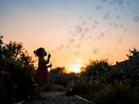 Young girl blowing soap bubbles at sunset in garden, silhouetted against golden sky with trees and fence, childhood wonder and outdoor play concept.