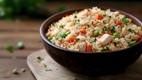 Homemade fried rice with chicken pieces, carrots, green peas and fresh herbs served in brown wooden bowl on rustic table, traditional Asian cuisine.
