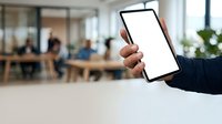 Hand holding blank tablet with white screen in modern office workspace with blurred colleagues in background, suitable for app mockups and business technology presentations.