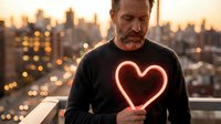 Middle-aged Caucasian man holding glowing red neon heart sign on urban rooftop at sunset with city skyline background for romantic Valentine's Day concept.