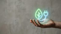 Hand holding glowing green leaf and electric plug icons representing sustainable energy, eco-friendly technology and environmental conservation concepts.