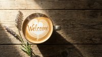 Steaming latte art coffee cup with Welcome text and heart on rustic wooden table with lavender, top view, warm cozy atmosphere.