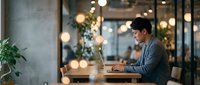 Young Asian businessman working on laptop in modern coworking space with warm lighting and industrial design elements for professional content.