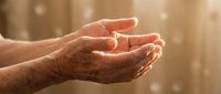 Elderly hands cupped together in gentle prayer or meditation pose with warm golden sunlight and soft bokeh background creating peaceful spiritual atmosphere.