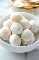 Fresh white snowball cookies dusted with powdered sugar served in ceramic bowl on marble surface, close-up view showing delicate texture and sweet coating.