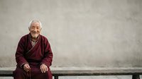 Senior Asian man with white hair wearing burgundy cardigan, sitting on bench against gray concrete wall, smiling at camera, suitable for aging, lifestyle, and portrait projects.