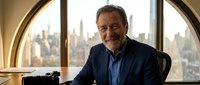 Senior male executive with gray beard and dark hair, appearing to be in his 60s, seated at desk in modern high-rise office with panoramic city skyline view through large arched windows, wearing navy blazer and blue shirt, professional business portrait.