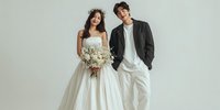 Young diverse couple in elegant wedding attire posing together on white background. Bride in white gown with bouquet, groom in black jacket and white pants.