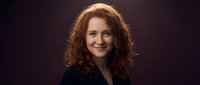 Professional portrait of confident young Caucasian woman with curly red hair smiling warmly against dark background for business and corporate use.