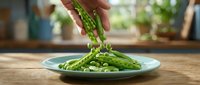 Fresh green asparagus spears being placed on a plate for healthy vegetable cooking and meal preparation in a bright kitchen setting.