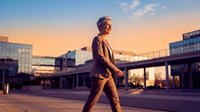 Senior man with gray hair, Caucasian ethnicity, wearing brown business suit, walking confidently across modern urban plaza at golden hour sunset, contemporary architecture and glass buildings in background.