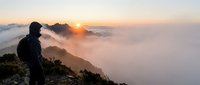 Hiker in dark jacket stands on mountain peak overlooking sea of clouds at sunrise, scenic landscape for travel and adventure content.