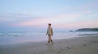 Young Asian man walking alone on serene beach at sunset with soft pink sky and gentle waves creating peaceful coastal atmosphere for wellness content.