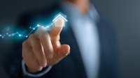 Professional in business suit pointing at glowing house icon with rising graph line, representing growth in real estate market and property investment trends.