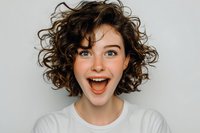 Young excited woman with curly brown hair showing surprised happy expression wearing white t-shirt on light background for lifestyle marketing.