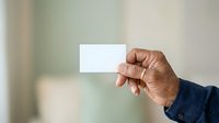 Middle-aged man with medium brown skin tone holding blank white business card in hand against soft blurred background, suitable for corporate branding and professional identity projects.