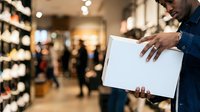 Man in denim shirt holding blank white tablet in retail store with shelves and customers in background, suitable for business and technology concepts.