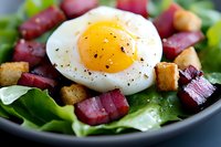 Fresh salad with sunny side up egg, crispy bacon bits, golden croutons, and mixed greens seasoned with black pepper served in dark bowl.