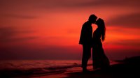 Romantic couple silhouette embracing at sunset beach with dramatic orange red sky and ocean waves creating intimate moment for love and relationship concepts.
