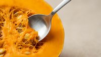 Fresh orange pumpkin cut in half showing fibrous interior with seeds being scooped out by metal spoon for cooking preparation and autumn recipes.
