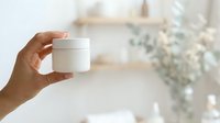 Hand holding white cosmetic jar with lid in bright bathroom setting, showcasing skincare product packaging for beauty and wellness marketing.