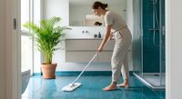 Young woman in beige uniform mopping modern bathroom floor with white mop, professional cleaning service in contemporary home interior with plants.