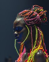African woman with neon-colored braids and face paint in profile view against dark background, dramatic lighting highlights vibrant pink, yellow and orange hair details.