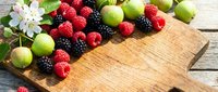 Fresh mixed berries and green apples arranged on rustic wooden cutting board with apple blossoms for healthy eating and cooking preparation.