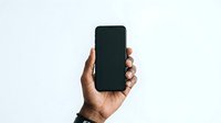 Person with medium-dark skin tone holding smartphone with blank black screen against white background for app mockup and digital marketing use.