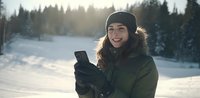 Young woman in winter clothing using smartphone outdoors with snowy landscape and forest in background during golden hour sunset.