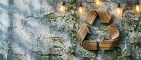 Wooden recycling symbol on textured concrete wall with warm string lights creating sustainable eco-friendly atmosphere for environmental awareness campaigns.