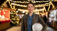 Professional Caucasian woman architect holding white hard hat at festive Christmas market with warm bokeh lights creating magical holiday atmosphere.