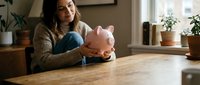 Young woman sitting at wooden table holding pink piggy bank planning personal finances and savings goals at home.