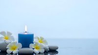Spa wellness scene with blue candle, white plumeria flowers and smooth stones on reflective surface for relaxation and meditation therapy.