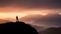Silhouette of person standing on mountain peak at sunset with dramatic orange and purple sky, misty valleys below, conveying achievement and contemplation for motivational and travel content.