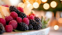 Fresh ripe raspberries and blackberries in white bowl with soft bokeh lights background, macro food photography for healthy eating and dessert concepts.