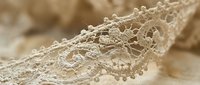 Delicate beige lace trim with intricate floral patterns and scalloped edges on neutral fabric background for sewing crafts and fashion design projects.
