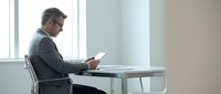 Middle-aged male professional, Caucasian ethnicity, wearing business casual attire and glasses, seated at a minimalist desk working on a laptop in a bright, modern office environment with natural light from windows.
