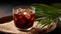Cold brew coffee with ice cubes in glass on wooden board with tropical palm leaves, dark moody background, selective focus, refreshing summer drink.