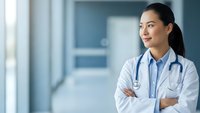 Confident Asian female doctor with stethoscope and crossed arms smiling in modern hospital corridor for healthcare and medical profession concepts.
