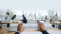 Businessman holding rolled architectural blueprints at desk with colleagues working in modern office overlooking city skyline, suitable for corporate and construction industry projects.